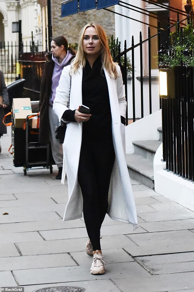 Kimberley Garner looks chic in a white trench coat and black roll neck top while glued to her phone